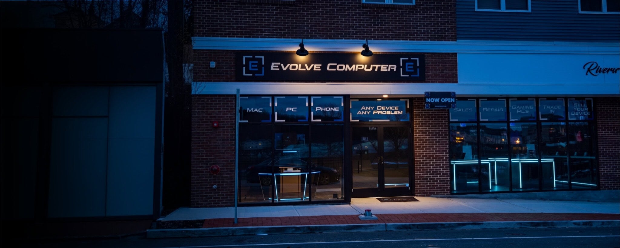 Night storefront of Evolve Computer with a brick facade, illuminated sign, and blue neon-lit windows advertising devices and services